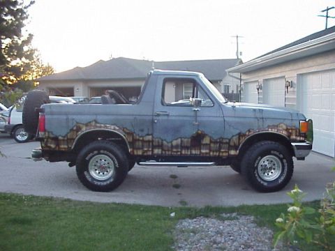 1989 Ford Bronco - Passenger-side profile of a lifted blue-gray Ford Bronco with the top removed and a roll bar exposed, parked in a residential driveway at d…