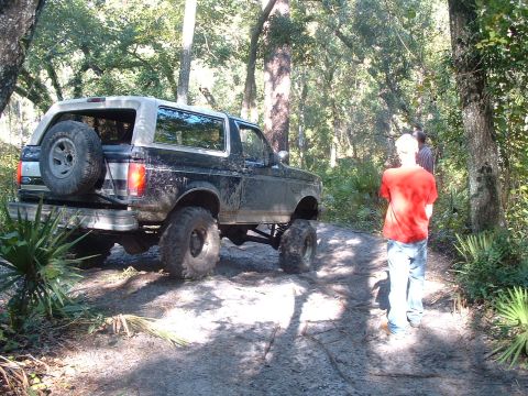 1995 Ford Bronco - A lifted Ford Bronco navigates a muddy trail surrounded by trees and foliage.