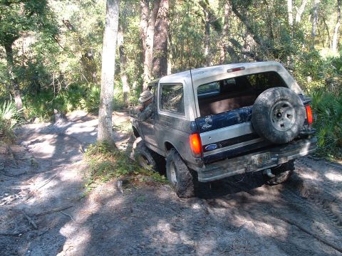 1995 Ford Bronco - A Ford Bronco navigates a muddy trail through a wooded area, showing its rear end and spare tire.