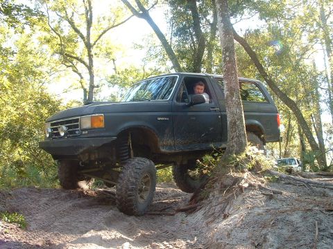 1995 Ford Bronco - A lifted black Ford Bronco navigates a challenging dirt path in a wooded area.
