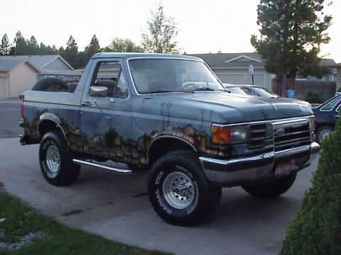 1989 Ford Bronco - A lifted full-size Ford Bronco with its hardtop removed, shot from the front three-quarter angle in a driveway. The body wears a custom air…