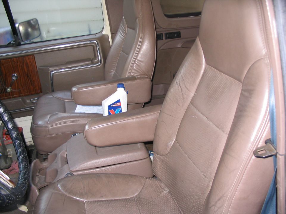 Interior shot of a vehicle's front seats, showing brown leather upholstery, armrests, and a bottle of oil on the passenger seat