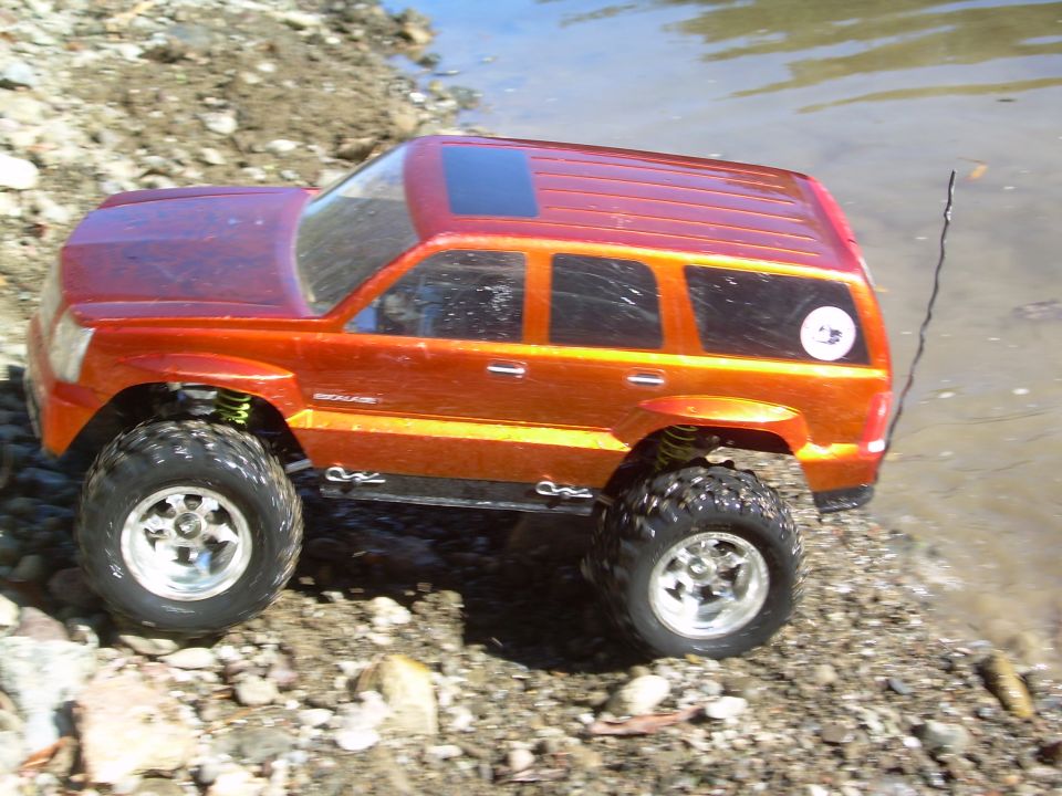 Orange RC SUV with large tires on rocky terrain near water