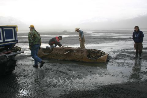 2003 Jeep TJ - A heavily rusted car body is partially submerged in mud, with several people observing it.