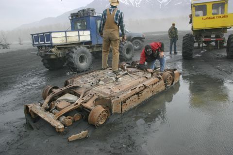 2003 Jeep TJ - A heavily rusted car chassis lies in muddy water, surrounded by large, off-road vehicles and people.