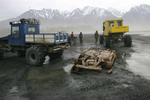 2003 Jeep TJ - Two custom off-road trucks are seen towing a rusted car frame in a muddy, mountainous landscape.
