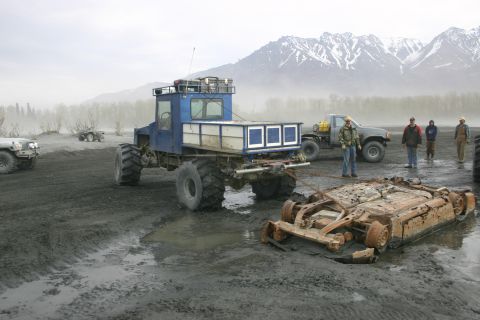2003 Jeep TJ - Several vehicles, including a heavily modified truck and a wrecked car, are present in a muddy, outdoor scene with mountains in the backgro…