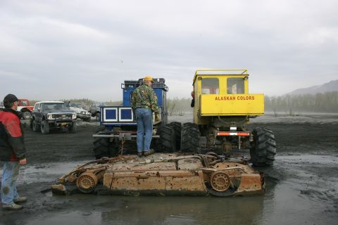 2003 Jeep TJ - A rusty car chassis lies in the mud, dwarfed by two large industrial vehicles and several onlookers.