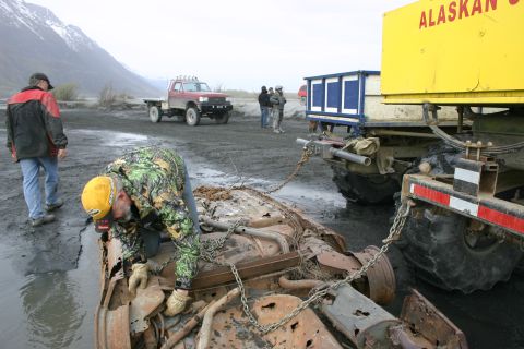 2003 Jeep TJ - A heavily damaged vehicle chassis is being towed by a large truck in a muddy, outdoor area.
