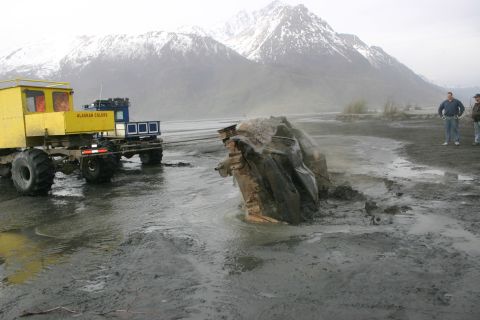2003 Jeep TJ - A yellow tow vehicle pulls heavily damaged wreckage from a muddy area, with snow-capped mountains visible in the distance.