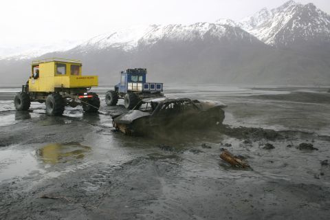 2003 Jeep TJ - Two large, all-terrain vehicles are pulling a heavily damaged car from a muddy, wet landscape with snow-capped mountains in the background.