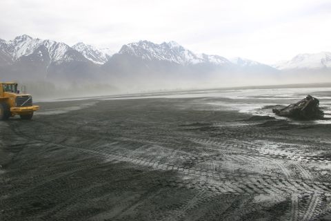 2003 Jeep TJ - A yellow front loader sits on a vast, muddy plain with tire tracks, under a hazy sky and distant snow-capped mountains.