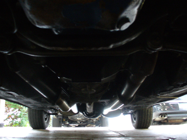 Undercarriage of a vehicle with dual exhaust pipes, showing the transmission and drive shaft. Another vehicle is partially visible in the background