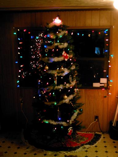 1986 Ford Bronco - A festive Christmas tree is adorned with colorful lights and ornaments, set against a wood-paneled wall and window.