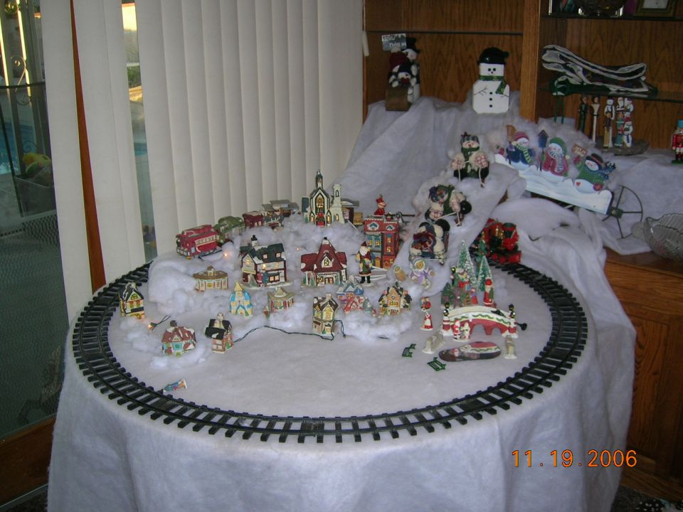 Christmas village display with miniature houses, trees, and a train track set up on a table covered in white fabric