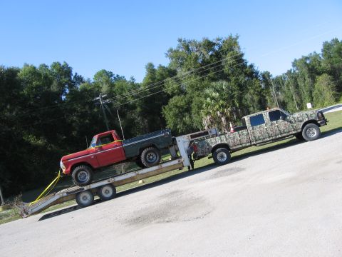 1974 Ford F-100 - A red and green pickup truck is transported on a trailer pulled by a camouflage truck on a sunny day.