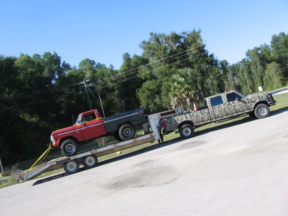 Red and green pickup truck loaded onto a trailer, which is being towed by a camouflage-painted crew cab truck