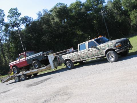 1974 Ford F-100 - A camouflage-wrapped truck tows a trailer carrying a two-tone pickup truck down a paved road.