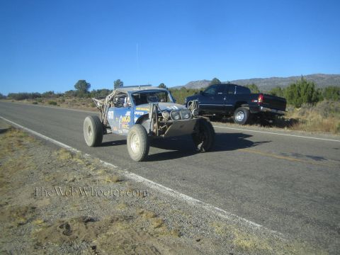 1990 Ford Bronco - gray and yellow tube-frame off-road race buggy travels a rural highway as a dark pickup sits parked roadside.