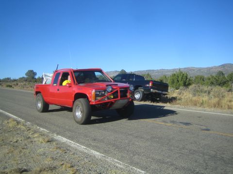 1990 Ford Bronco - red extended-cab pickup with a brush guard and off-road tires drives a rural highway near a parked dark truck.