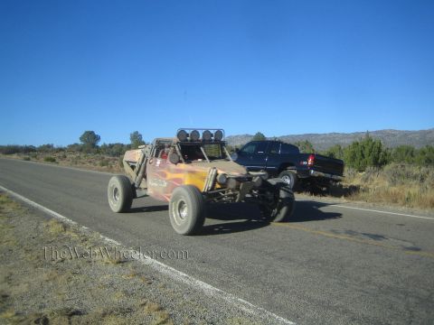 1990 Ford Bronco - dark flame-painted tube-frame off-road race buggy drives a rural highway as a dark pickup sits on the shoulder.