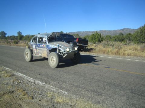 1990 Ford Bronco - gray Volkswagen Beetle-bodied off-road race car with headlights on drives a rural highway past a parked pickup.