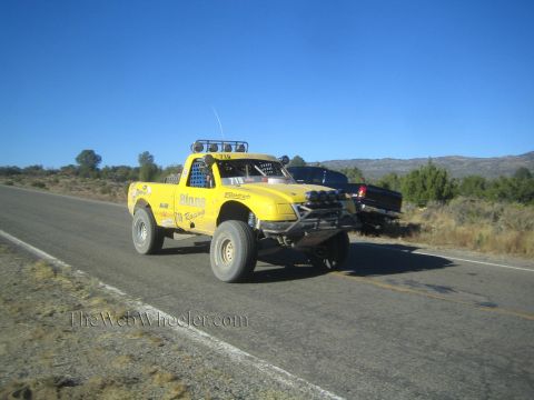1990 Ford Bronco - yellow off-road race truck marked Pinos Racing drives a rural highway as a dark pickup sits parked on the shoulder.
