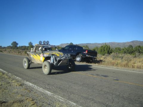 1990 Ford Bronco - black and yellow tube-frame off-road race buggy travels a rural highway as a dark pickup sits parked roadside.