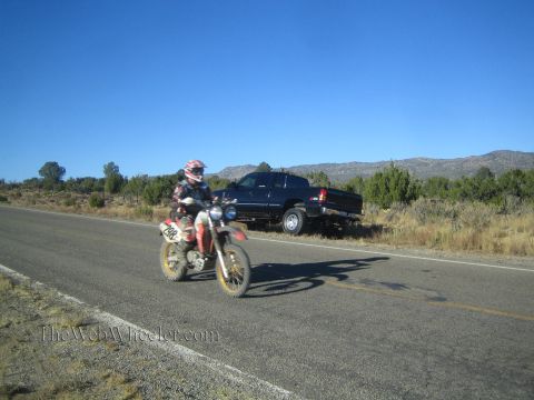 1990 Ford Bronco - helmeted rider on a red Honda dirt bike rides along a rural highway as a dark pickup sits parked behind.