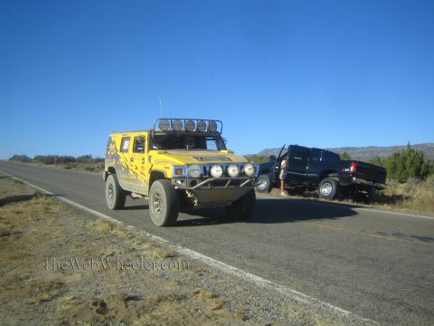 1990 Ford Bronco - yellow Hummer H2 with a roof-mounted light bar and brush guard drives a rural highway past a parked dark pickup.