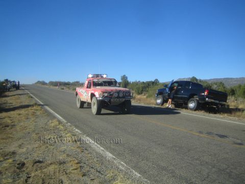 1990 Ford Bronco - red off-road race truck drives a rural highway while a person stands beside a parked dark pickup on the shoulder.