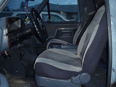 1989 Ford Bronco - The front interior of a truck with two-tone gray-and-black cloth bucket seats, a lace-up steering-wheel cover, a woodgrain-accented dash, a…