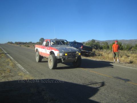 1990 Ford Bronco - red, white and blue off-road race truck drives a rural highway as a man in an orange shirt watches from the shoulder.