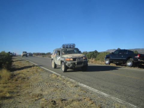 1990 Ford Bronco - olive-green Toyota FJ Cruiser with a roof light bar drives a rural highway near parked vehicles and spectators.