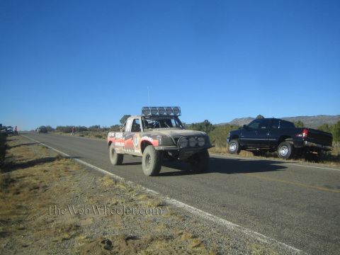 1990 Ford Bronco - tan and red off-road race truck drives a rural highway as a dark pickup sits parked on the shoulder.