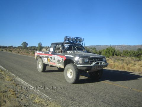 1990 Ford Bronco - black off-road race truck with sponsor decals and a roof light bar drives a rural highway in hilly terrain.