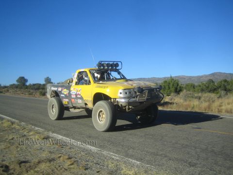 1990 Ford Bronco - yellow off-road race truck covered in sponsor decals, fitted with a roof-mounted light bar, drives down a paved desert highway.