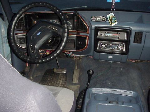 1989 Ford Bronco - Over-the-shoulder interior shot of a blue-grey Ford truck dashboard, showing a laced-on steering wheel cover, woodgrain instrument bezel wi…