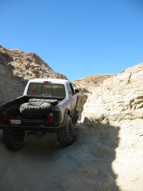 1997 Ford Ranger - A white pickup truck navigates a narrow dirt path between rocky canyon walls, carrying a spare tire in its bed.