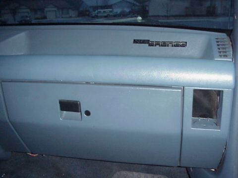 1989 Ford Bronco - Passenger side of a blue-gray Ford dashboard showing the closed glovebox door with its latch handle and the black 4x4 Bronco emblem badge o…