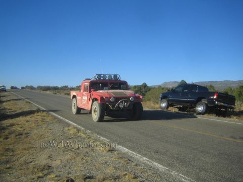1990 Ford Bronco - red Hummer H1 with a roof light bar travels along a desert highway while a dark pickup truck sits pulled off on the roadside.