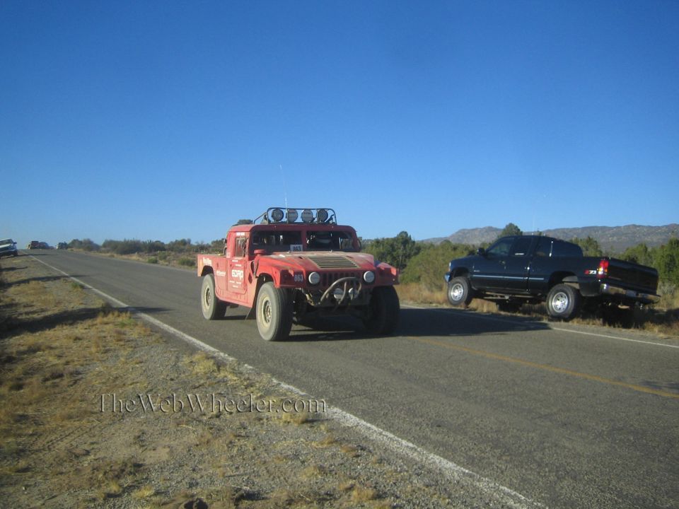 Red Hummer H1 on a desert highway with a dark pickup pulled off to the side