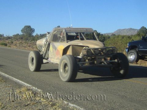 1990 Ford Bronco - tan, dusty single-seat off-road race buggy with large knobby tires drives along a desert highway with hills in the background.