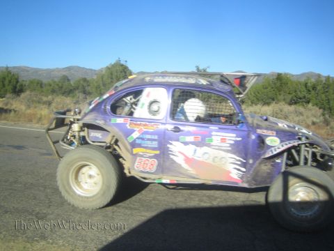 1990 Ford Bronco - purple Baja Bug built from a Volkswagen Beetle, numbered 568 and covered in decals, races along a paved desert road.
