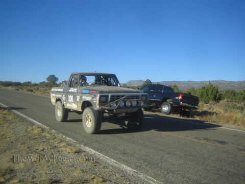 1990 Ford Bronco - vintage olive-drab Ford off-road race truck numbered 300 with front light pods drives a desert highway as a dark pickup waits roadside.
