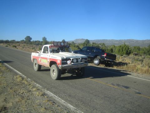 1990 Ford Bronco - white and red 1970s square-body Chevrolet race pickup with a roof light bar drives a desert highway beside a dark parked pickup.