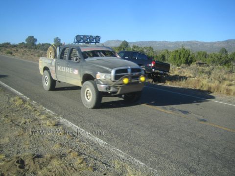 1990 Ford Bronco - Dodge Ram crew-cab prerunner labeled KORE, fitted with a roof light bar and yellow fog lights, drives a desert highway.