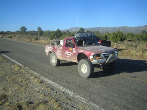 1990 Ford Bronco - dark red single-cab prerunner race truck with a roof light bar and sponsor decals drives down a paved desert highway.