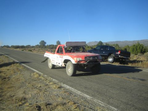 1990 Ford Bronco - white and red prerunner race truck with a roof light bar travels a desert highway with a dark pickup pulled off to the side.