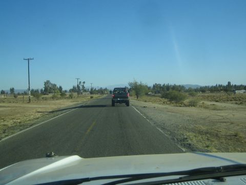 1990 Ford Bronco - Seen through a vehicle windshield, a dark pickup truck travels ahead on a rural paved road lined with power poles and dry brush.
